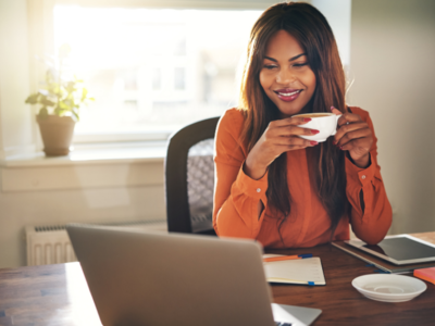 A woman enjoys a coffee at her desk as she watches a webinar on her laptop