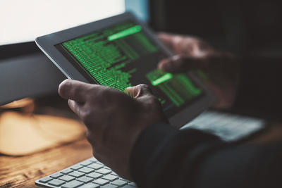 A man holds a tablet, whilst sitting in front of a computer