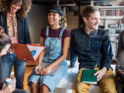A group of people are sitting and standing together, smiling and holding documents, books, and a laptop.