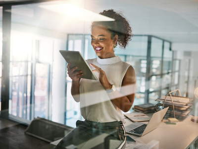 A smiling person in a modern office reads a document on a tablet while standing beside an open laptop and a desk with folders, with glass walls and warm lighting in the background.